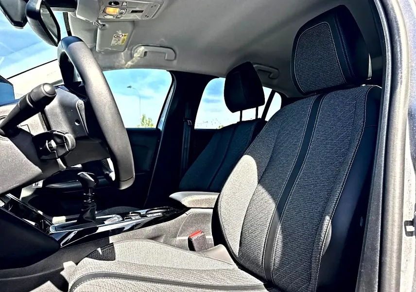 Intérieur de la Peugeot 208 1.2 100 Allure 2025, vue côté conducteur, sièges tissu gris et console centrale noire brillante.