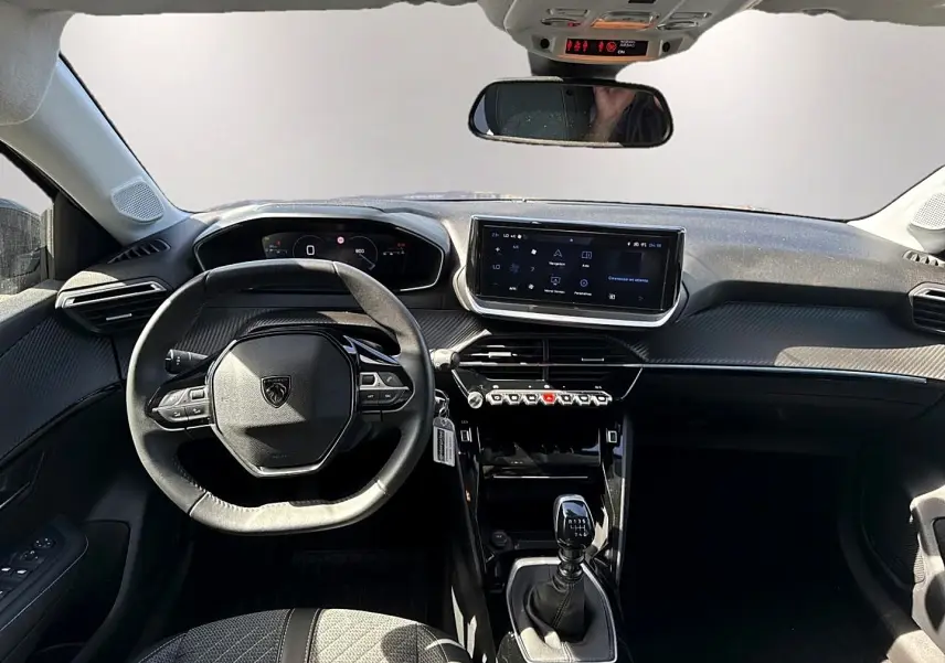Vue intérieure avant de la Peugeot 208 1.2 100 Allure 2025, tableau de bord noir avec écran tactile et volant compact.
