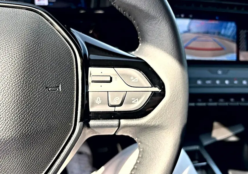 Gros plan sur le volant cuir noir de la Peugeot 308 2025 avec commandes audio et écran de caméra de recul en arrière-plan.
