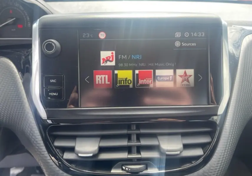 Vue rapprochée de l’écran tactile central de la Peugeot 208 blanc perle, affichant les stations radio avec commandes autour.