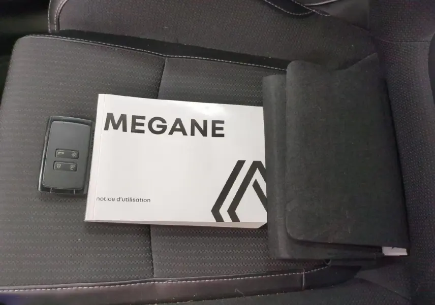 Notice d'utilisation Mégane et clé Renault posées sur un siège avant en tissu noir avec surpiqûres grises.