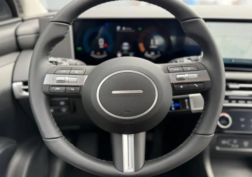 Vue rapprochée du volant noir du Hyundai Tucson 2026 avec commandes intégrées et tableau de bord numérique flou en arrière-plan.