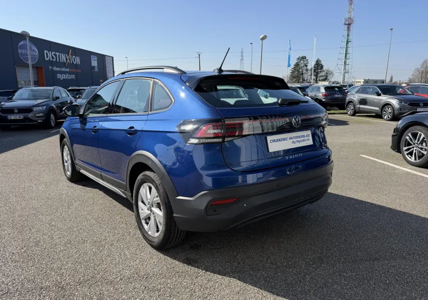 Vue 3/4 arrière droite d'un Volkswagen Taigo bleu métallisé avec barres de toit et feux arrière LED.