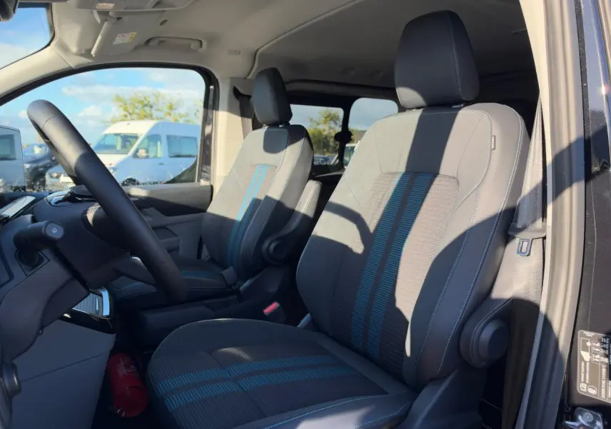 Vue intérieure côté conducteur du Ford Transit Fourgon 2026, sièges sport gris avec bandes bleues et volant noir.