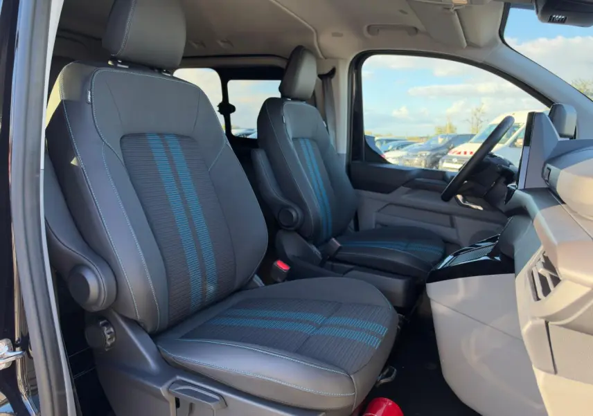 Vue intérieure côté conducteur du Ford Transit Fourgon 2026, sièges sport noirs avec bandes bleues et tableau de bord gris.