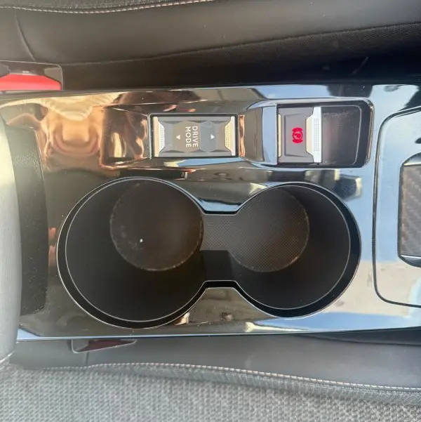 Vue plongeante sur la console centrale noire de la Peugeot 208 Hybrid 110 e-DCS6 Allure 2024 avec porte-gobelets et boutons Drive Mode et frein à main.