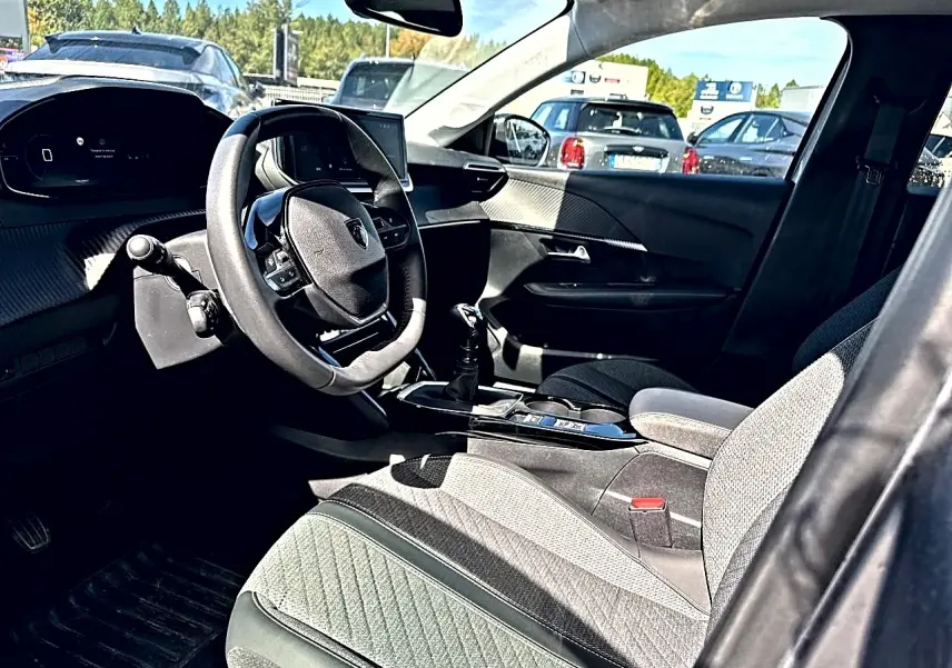 Vue intérieure côté conducteur de la Peugeot 208 1.2 100 Allure 2025 avec volant compact et sellerie tissu gris.