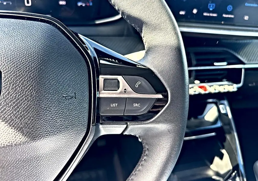 Gros plan sur le volant noir de la Peugeot 208 1.2 100 Allure avec commandes audio intégrées, tableau de bord numérique en arrière-plan.