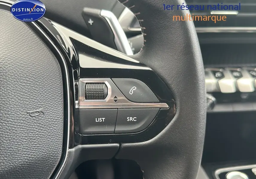Gros plan sur les commandes gauche du volant cuir noir du Peugeot 5008 2024 avec tableau de bord en arrière-plan flou.