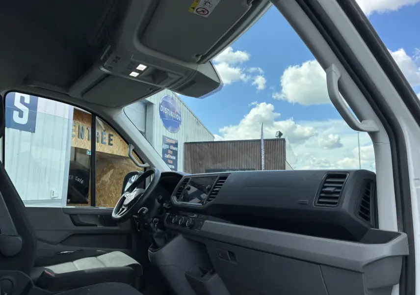 Vue intérieure côté conducteur du Volkswagen Crafter blanc, montrant tableau de bord et volant multifonctions sous ciel bleu.