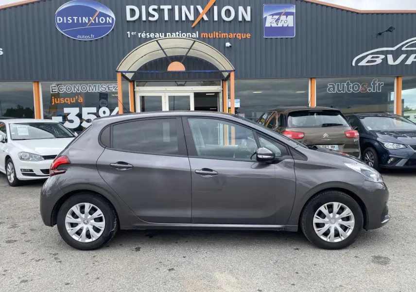 Profil côté gauche d'une Peugeot 208 gris métallisé de 2018, compacte et cinq portes, stationnée devant un concessionnaire.