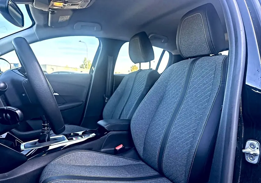 Vue intérieure côté conducteur de la Peugeot 208 noire 2025, mettant en valeur les sièges en tissu gris et le volant noir.