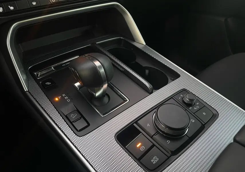 Gros plan sur la console centrale du Mazda CX-60 2024, avec levier de vitesses et commandes noires sur finition argentée texturée.