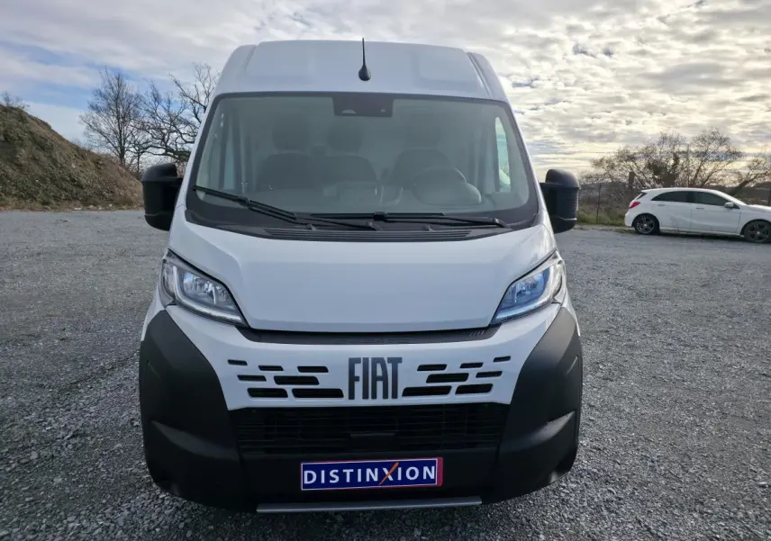 Vue de face d'un FIAT Ducato Fourgon blanc avec pare-chocs noir et logo FIAT bien visible sur un terrain extérieur.