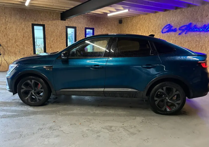 Vue de profil côté gauche du Renault Arkana bleu 1.3 TCE 140 RS-LINE avec jantes noires et détails rouges, stationné en intérieur.