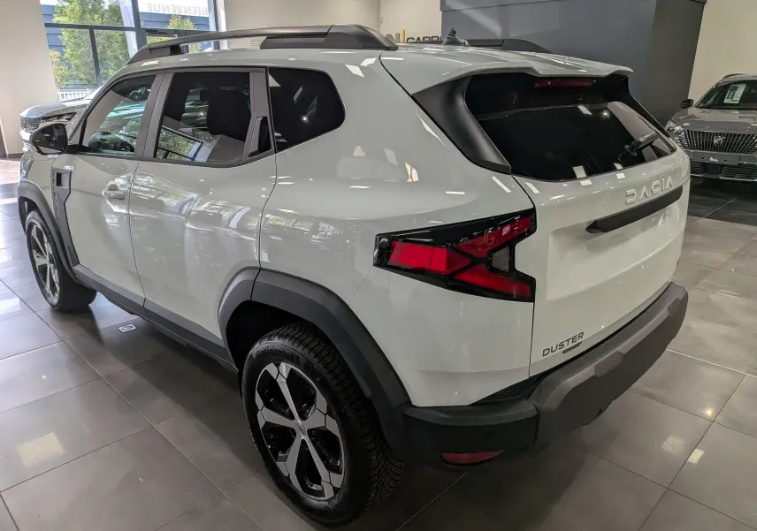 Vue 3/4 arrière droite du Dacia Duster blanc Glacier avec jantes bi-ton et barres de toit noires en showroom.