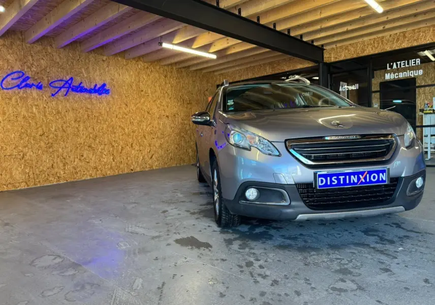 Peugeot 2008 gris en 3/4 avant droit dans un garage avec mur en bois et enseigne lumineuse bleue.