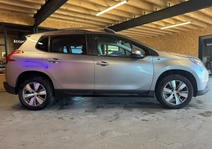 Peugeot 2008 gris vue de profil côté gauche, SUV compact avec jantes alliage et barres de toit en intérieur bois.