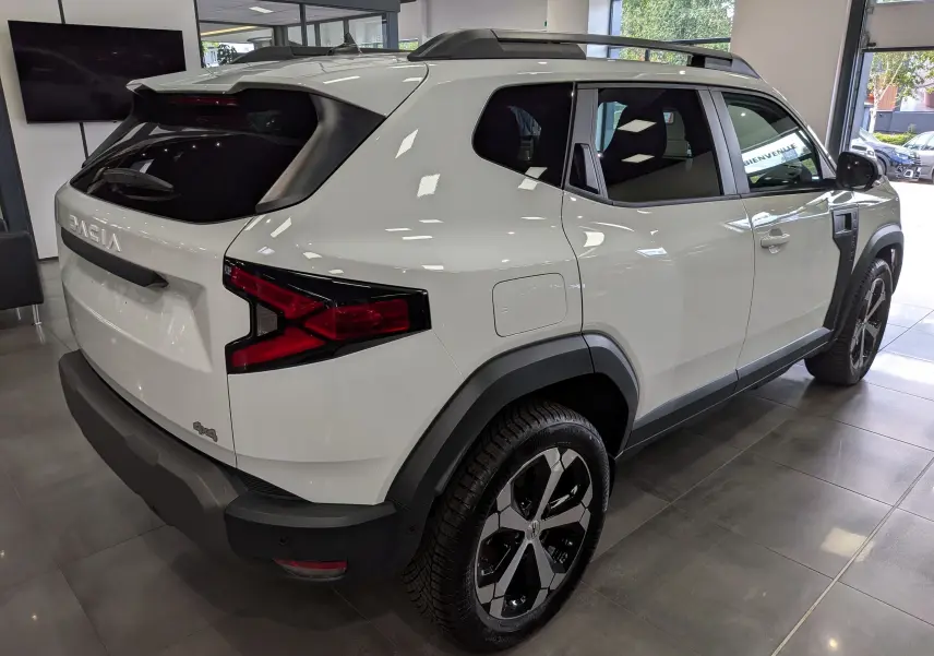 Vue 3/4 arrière droite du Dacia Duster Hybrid-G blanc Glacier avec jantes bi-ton et protections noires contrastantes.