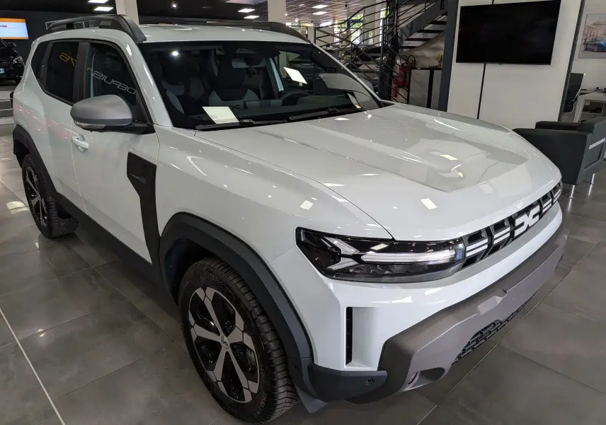 Vue 3/4 avant droite du Dacia Duster Hybrid-G blanc Glacier avec jantes bi-ton et protections noires contrastantes.