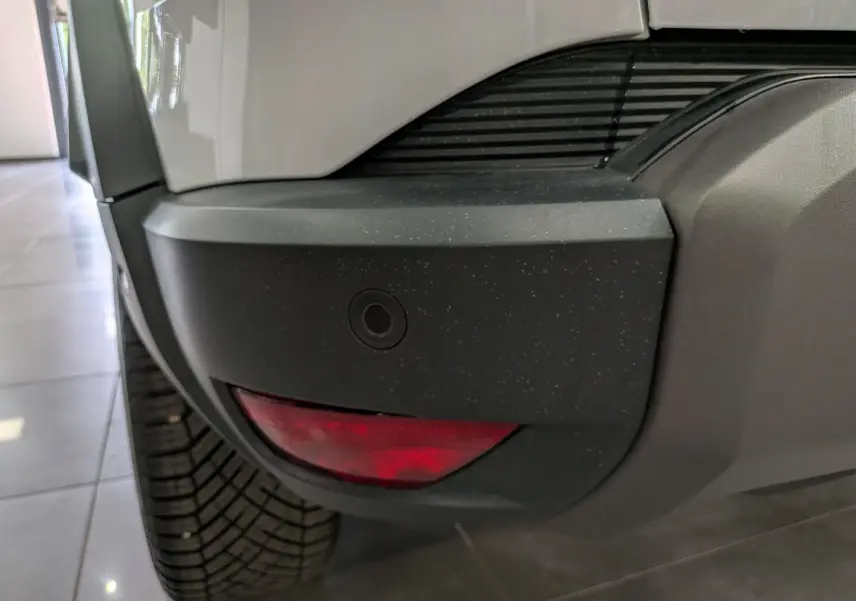 Gros plan sur l'angle arrière gauche du Dacia Duster blanc Glacier, montrant le pare-chocs noir avec feu rouge et capteur de recul.