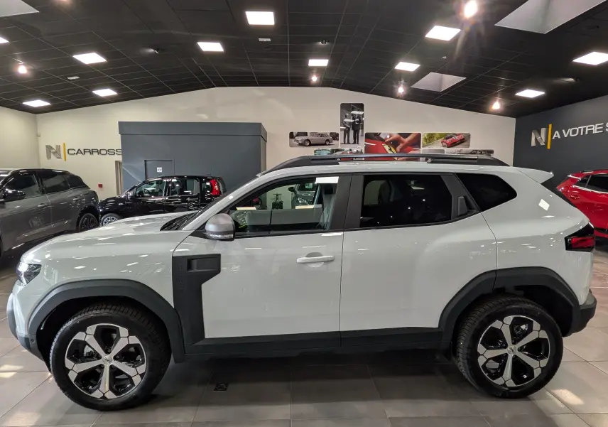 Vue de profil côté gauche du Dacia Duster Hybrid-G 150 CH 4x4 blanc glacier avec jantes bi-ton et protections noires.