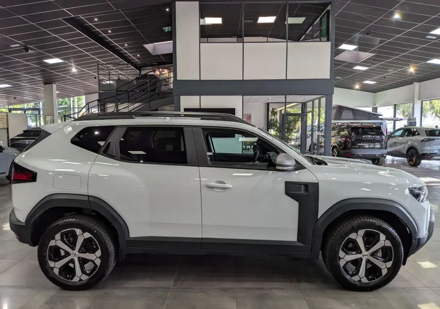 Vue latérale droite d'un Dacia Duster Hybrid-G blanc Glacier 2026 avec jantes bi-ton et protections noires sur les ailes.