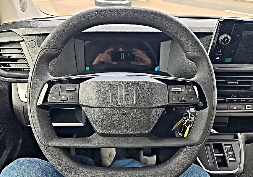 Vue intérieure centrée sur le volant noir FIAT avec commandes intégrées et tableau de bord numérique en arrière-plan.