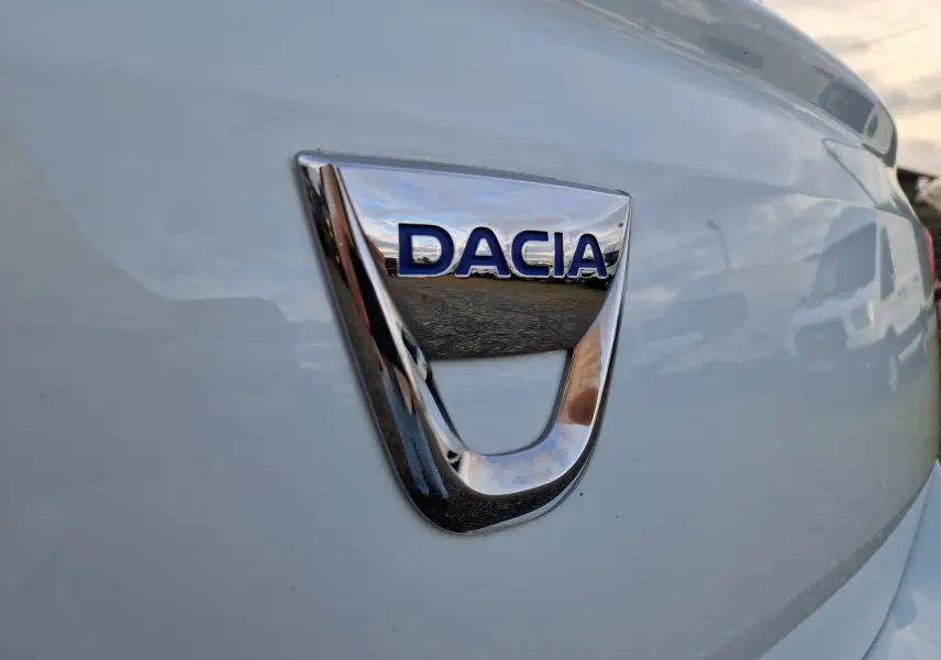 Gros plan sur le logo chromé DACIA à l'arrière d'une Dacia Sandero blanche, vue de trois quarts arrière droit.