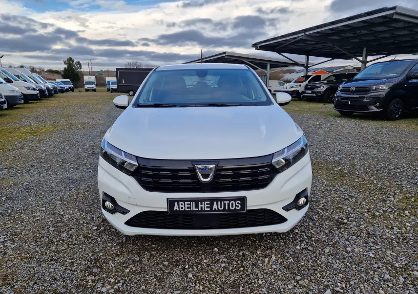 Vue frontale d'une Dacia Sandero blanche 2022 avec calandre noire et phares LED allumés sur un parking.