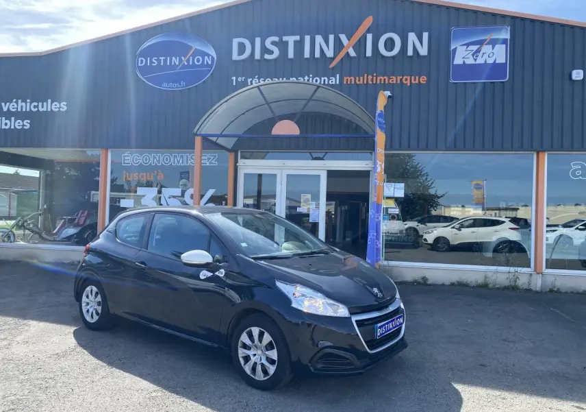 Peugeot 208 gris foncé vue 3/4 avant droit, avec enjoliveurs 15 pouces et rétroviseurs blancs devant un garage Distinxion.