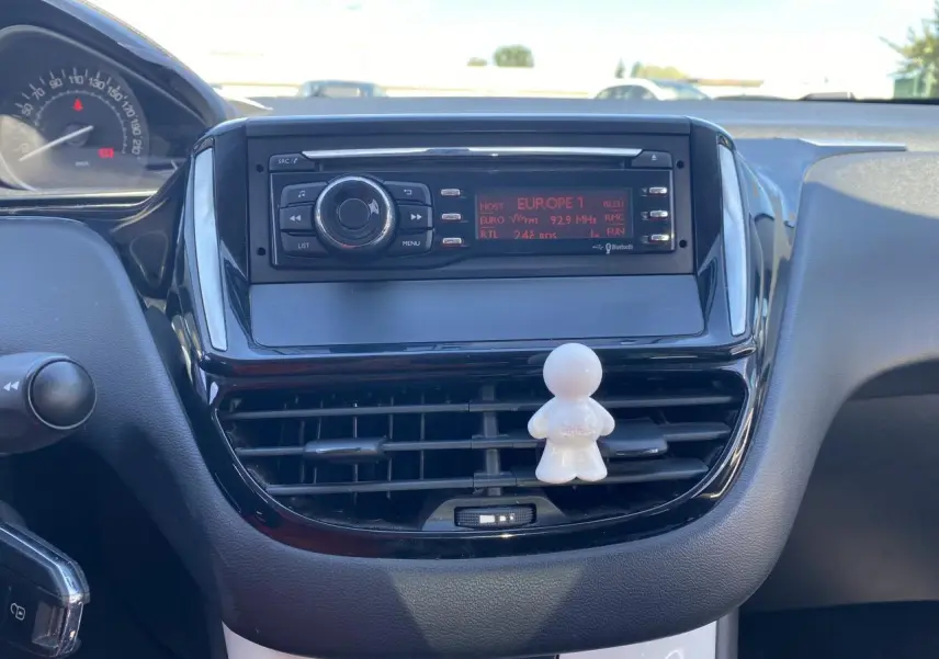 Vue rapprochée du tableau de bord de la Peugeot 208 gris foncé, avec autoradio et ventilation centrale décorée d’un petit personnage blanc.