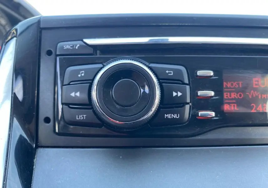 Gros plan sur l'autoradio avec molette centrale et écran rouge affichant les stations, intérieur Peugeot 208 gris foncé.