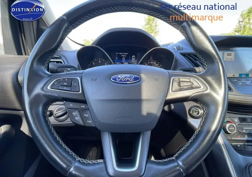 Vue intérieure centrée sur le volant cuir noir du Ford Kuga blanc avec tableau de bord et écran tactile visible.