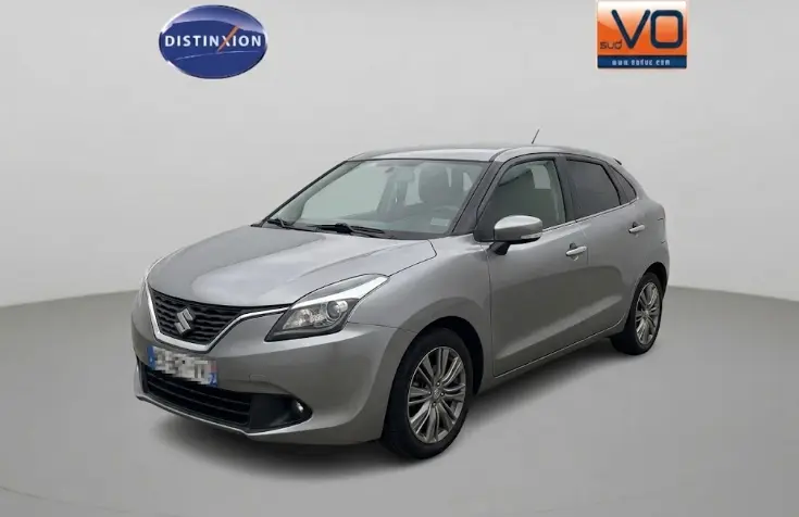 Suzuki Baleno gris clair vue 3/4 avant droit, jantes alliage 16 pouces et vitres surteintées visibles.