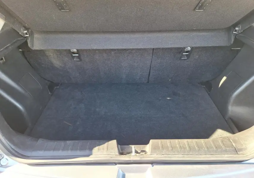 Coffre ouvert d'une Suzuki Baleno gris clair, montrant l'espace de rangement intérieur et la moquette noire.