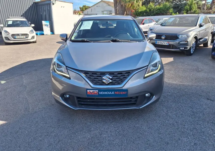 Vue avant d'une Suzuki Baleno gris clair 2017 avec phares allumés et plaque "Véhicule récent" visible.