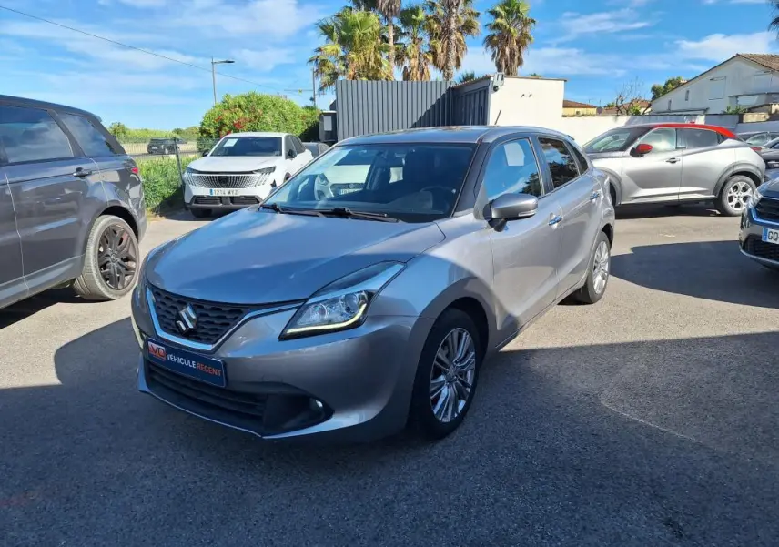 Suzuki Baleno gris clair vue 3/4 avant droit, jantes alliage 16 pouces et phares allumés en extérieur.