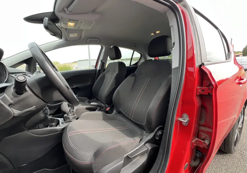 Vue intérieure côté conducteur d'une Opel Corsa rouge avec sièges tissu noirs à surpiqûres rouges et volant cuir.