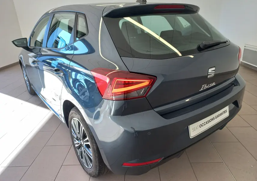 Vue 3/4 arrière droite d'une SEAT Ibiza 2025 gris magnétique avec feux arrière allumés dans un showroom.