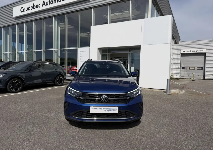 Vue de face du Volkswagen Taigo bleu métallisé garé devant un showroom Caudebec Automobiles par temps clair.