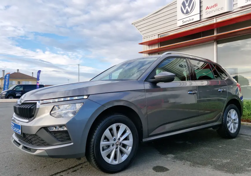 Skoda Kamiq gris graphite métal en 3/4 avant droit, avec calandre noire et jantes alliage visibles devant un garage VW.