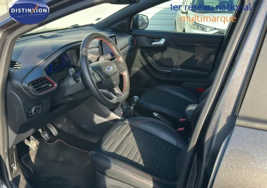 Vue intérieure côté conducteur d'un Ford Puma gris foncé, montrant sièges noirs avec surpiqûres rouges et tableau de bord moderne.