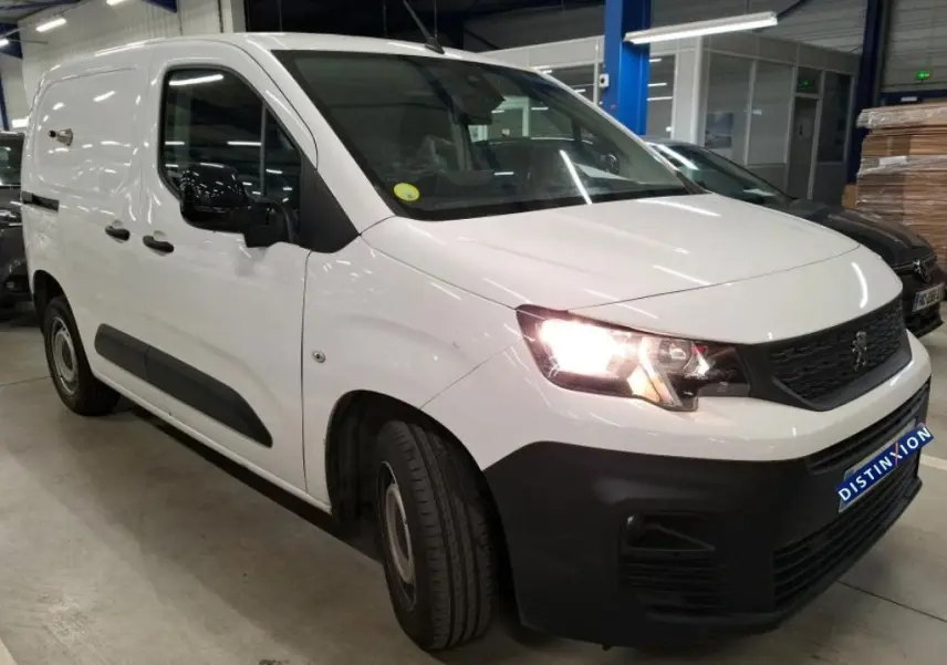 Vue 3/4 avant droit d'un Peugeot Partner Fourgon blanc 2022 avec phares allumés et protections noires sur les bas de caisse.