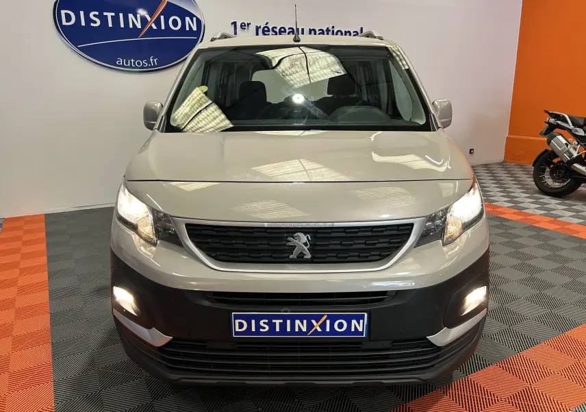 Vue frontale d'un Peugeot Rifter BlueHDi 100 Active gris sable avec phares allumés en intérieur showroom.