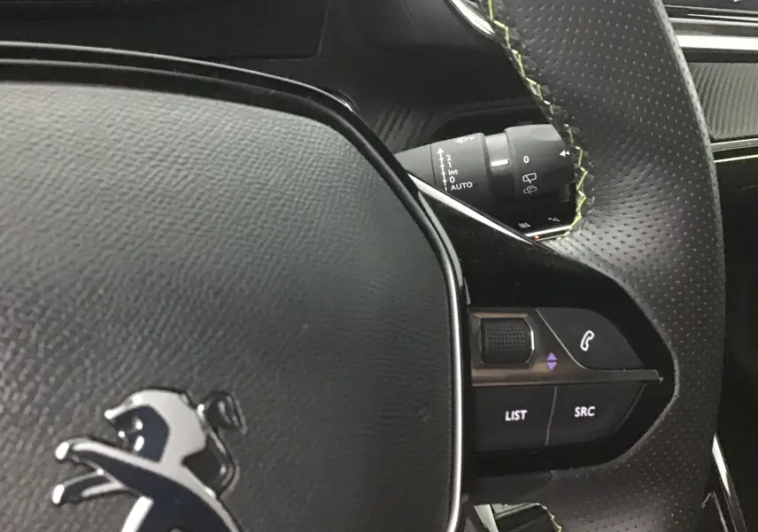 Gros plan sur le volant cuir perforé noir avec surpiqûres vertes du Peugeot 2008 électrique gris Artense, montrant les commandes audio.