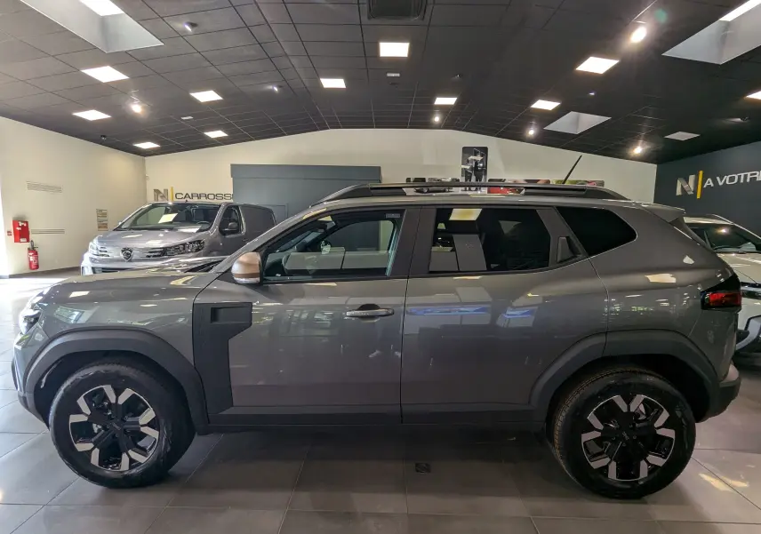 Profil côté gauche du Dacia Duster 2025 gris schiste Mild Hybrid 130 CH 4x2 EXTRÊME en showroom avec jantes noires et toit noir.