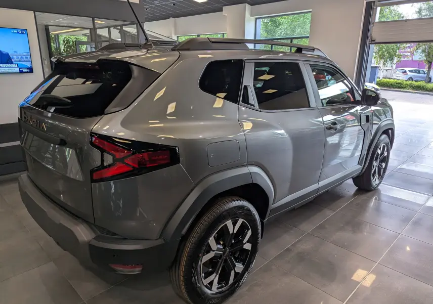 Vue 3/4 arrière droite du Dacia Duster 2025 gris schiste avec jantes noires et toit équipé de barres de toit.