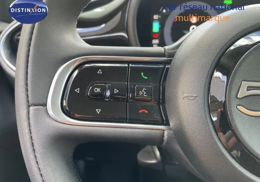 Gros plan sur le volant cuir noir de la Fiat 500 électrique 2023, avec commandes multifonctions et tableau de bord digital en arrière-plan.