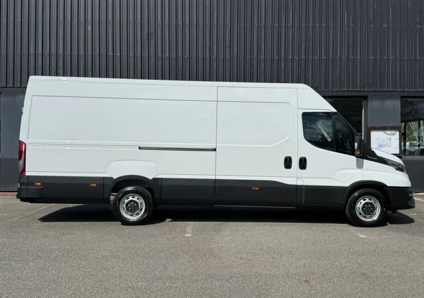 Fourgon Iveco Daily blanc vu de profil côté gauche, avec bas de caisse noir et roues acier, stationné devant un mur sombre.